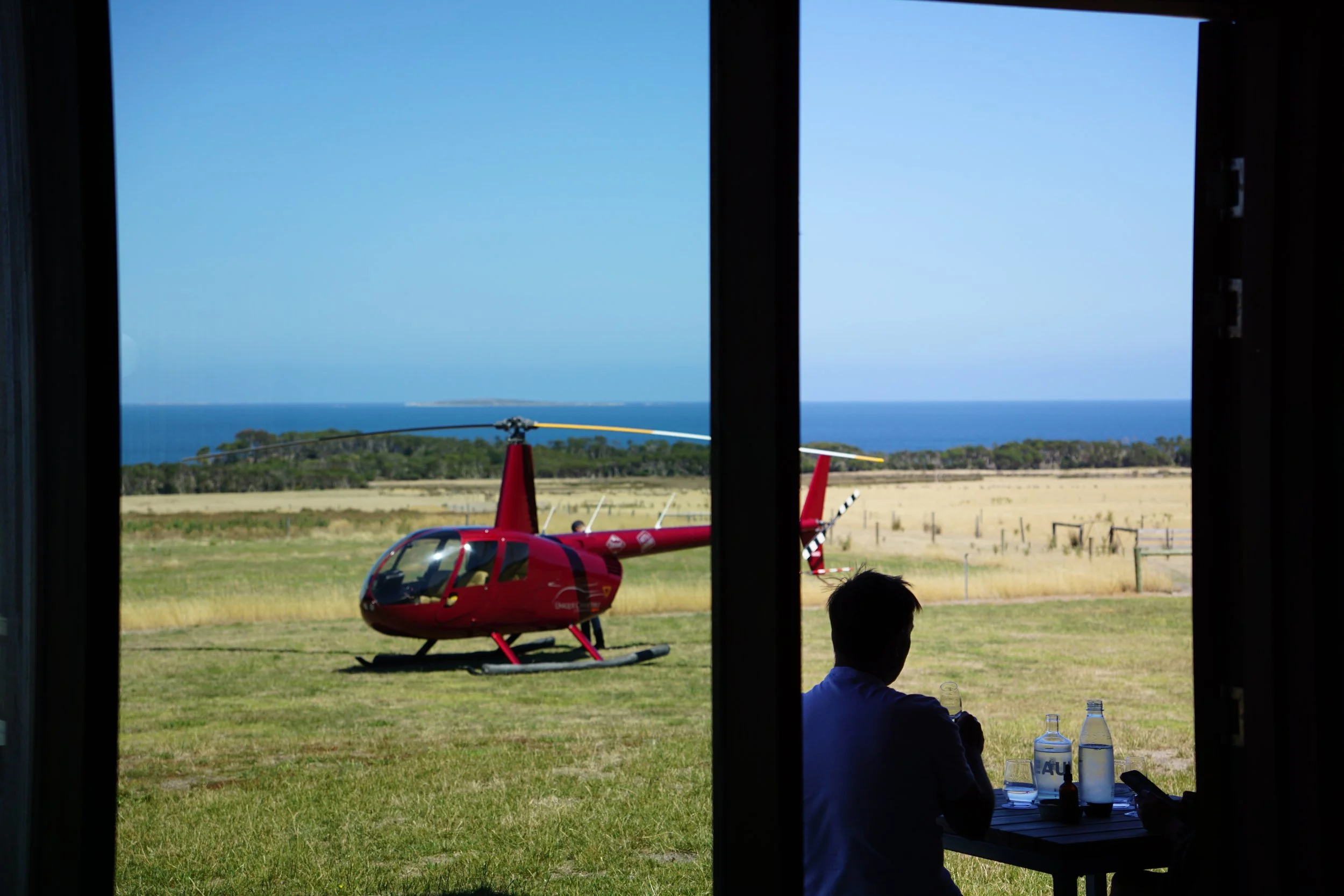 View through a window showing a small red helicopter parked on a grassy field with a person sitting at a table with water bottles in the foreground. In the background, there are trees, a fence, and the ocean under a clear sky.