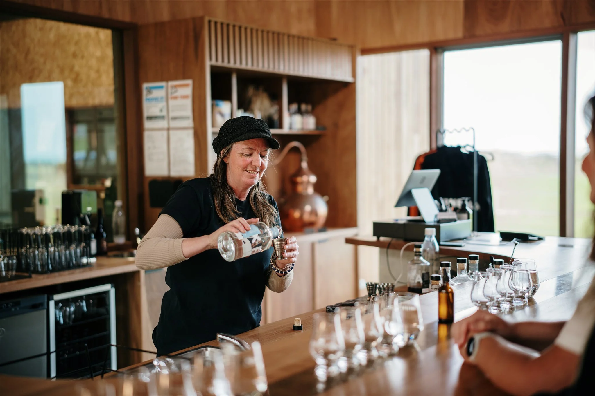 A woman with long hair wearing a black beanie and black shirt pours liquid from a glass bottle into a metal measuring cup at a rustic wooden bar with various bottles and glasses, outdoor scenery visible through large windows.