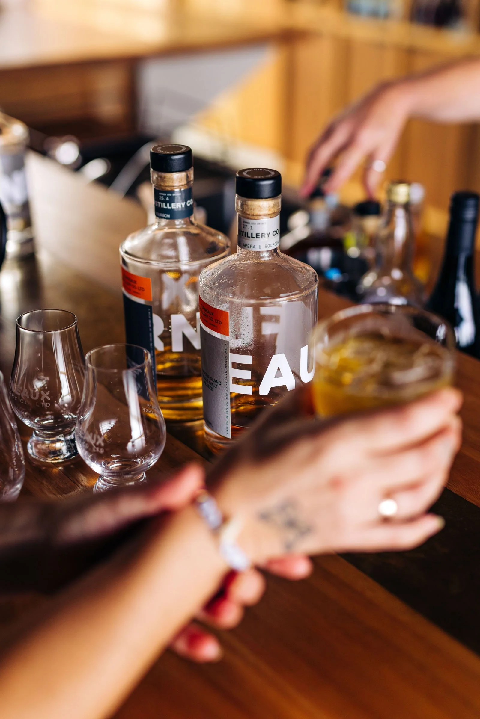 Close-up of two bottles of alcohol on a wooden bar, with different glasses nearby, and a person reaching for a bottle in the background.