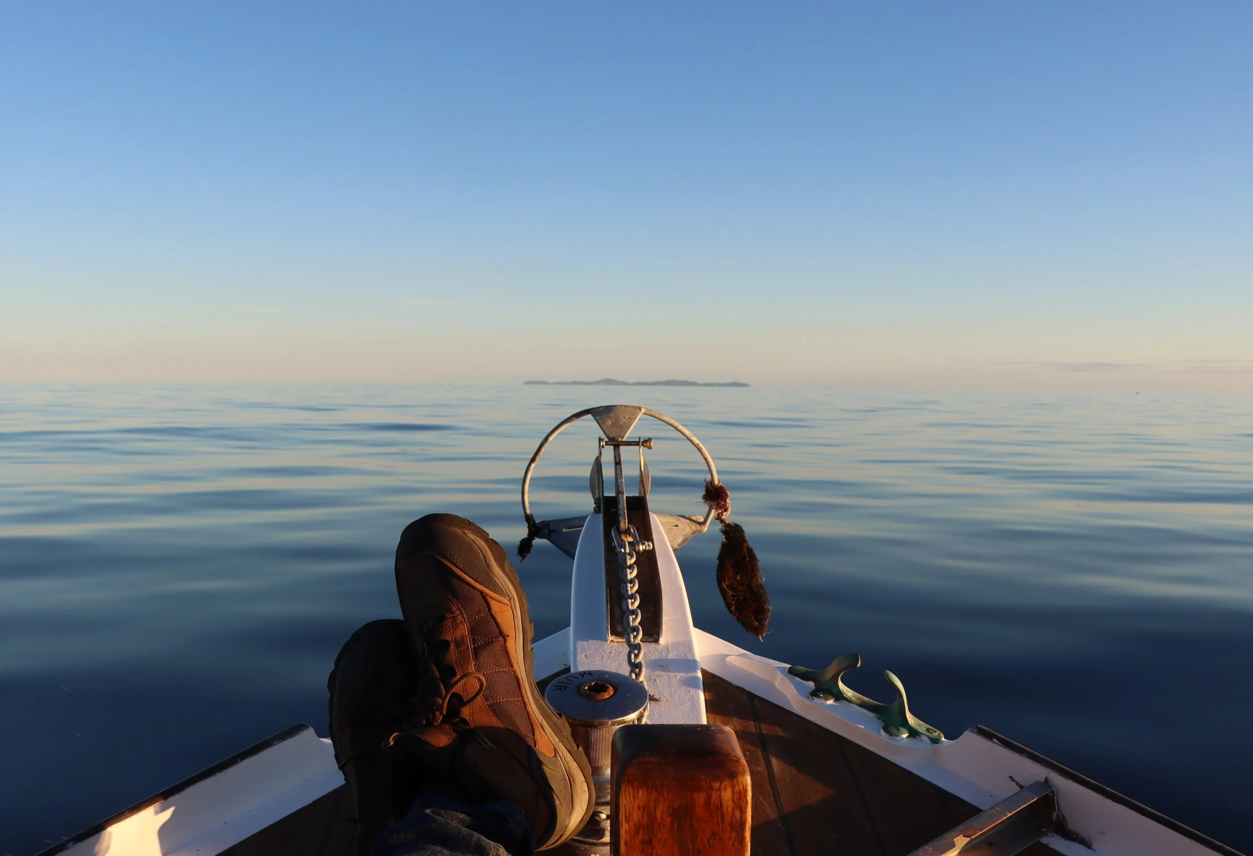 Person wearing brown shoes sitting on a kayak or small boat, with feet up, on calm water. The view shows a distant landmass and a clear blue sky.