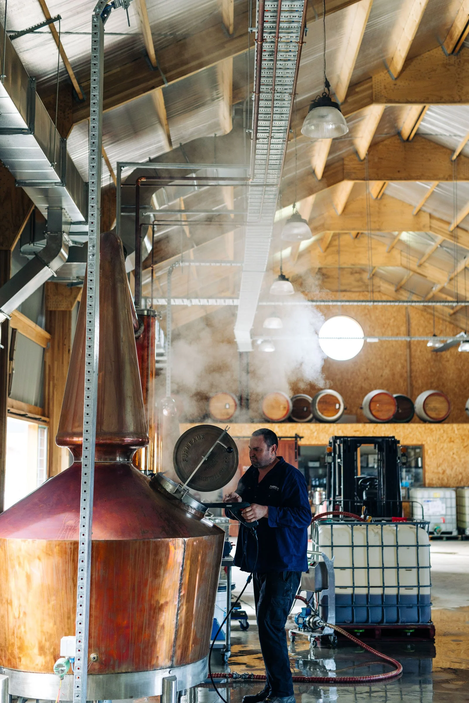 A man working in a brewery with a large copper brewing kettle, pipes, and filtration equipment. Shelves of barrels and industrial lighting are visible in the background.