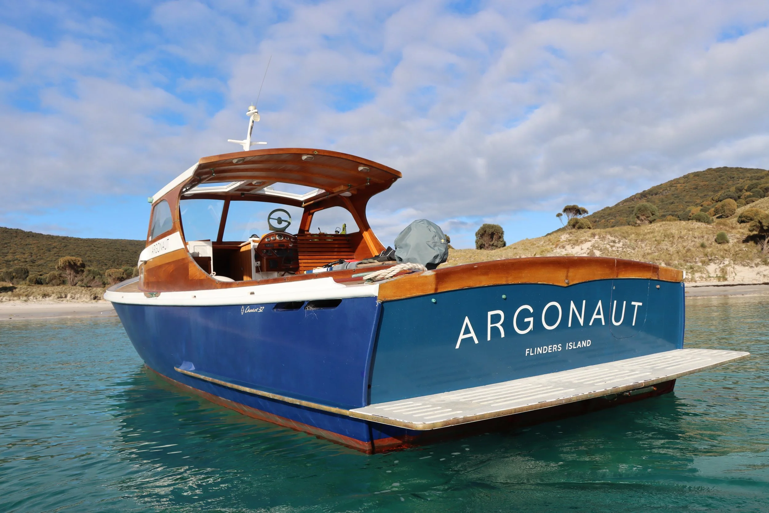 A wooden boat named Argonaut floating on clear water near a shoreline with hills and trees in the background. The boat has a covered area with windows and a steering wheel inside.