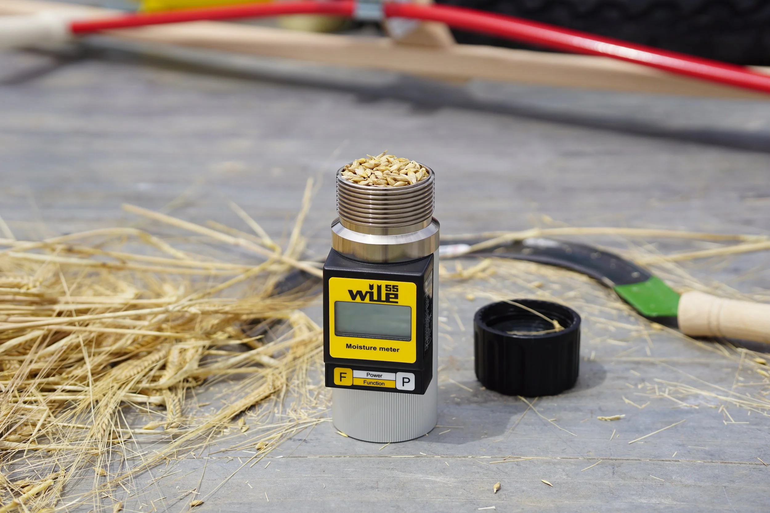 A moisture meter with a digital display and a stack of sunflower seeds on top, surrounded by straw and various tools in a work environment.