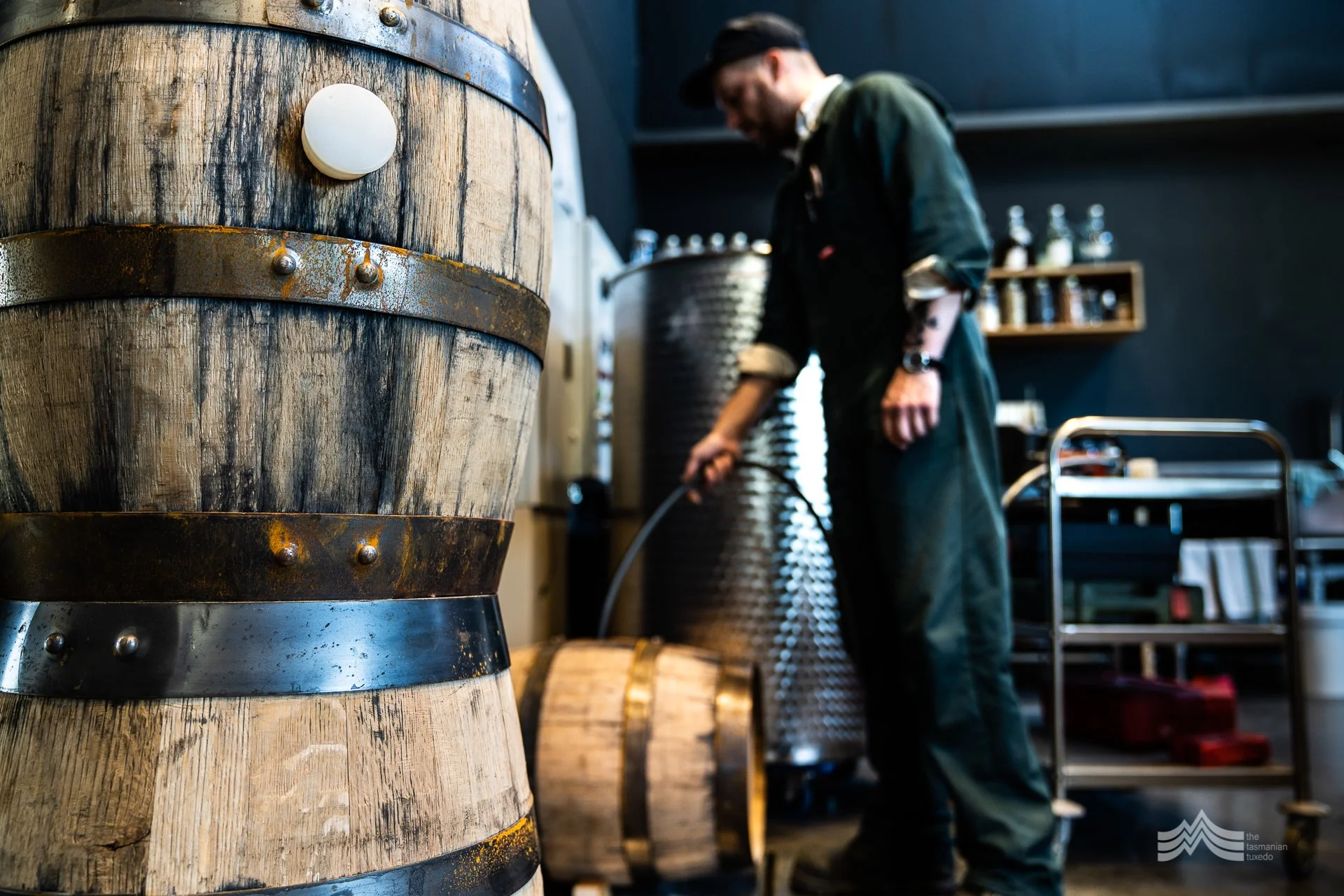 A man in a green work overall and black cap working in a distillery or winery with large wooden and metal fermentation or storage tanks in the background, focused on his task.