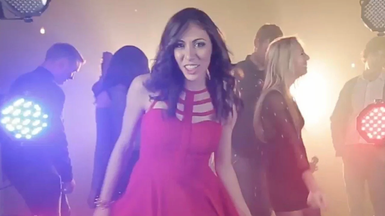 Group of people at a dance party, with a woman in a red dress smiling at the camera, colorful lights, and foggy atmosphere.