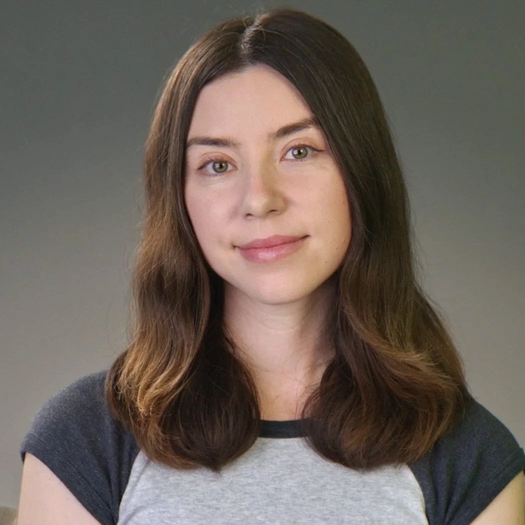 Portrait of a young woman with shoulder-length brown hair and light skin, wearing a gray T-shirt with black trim, looking at the camera with a slight smile.