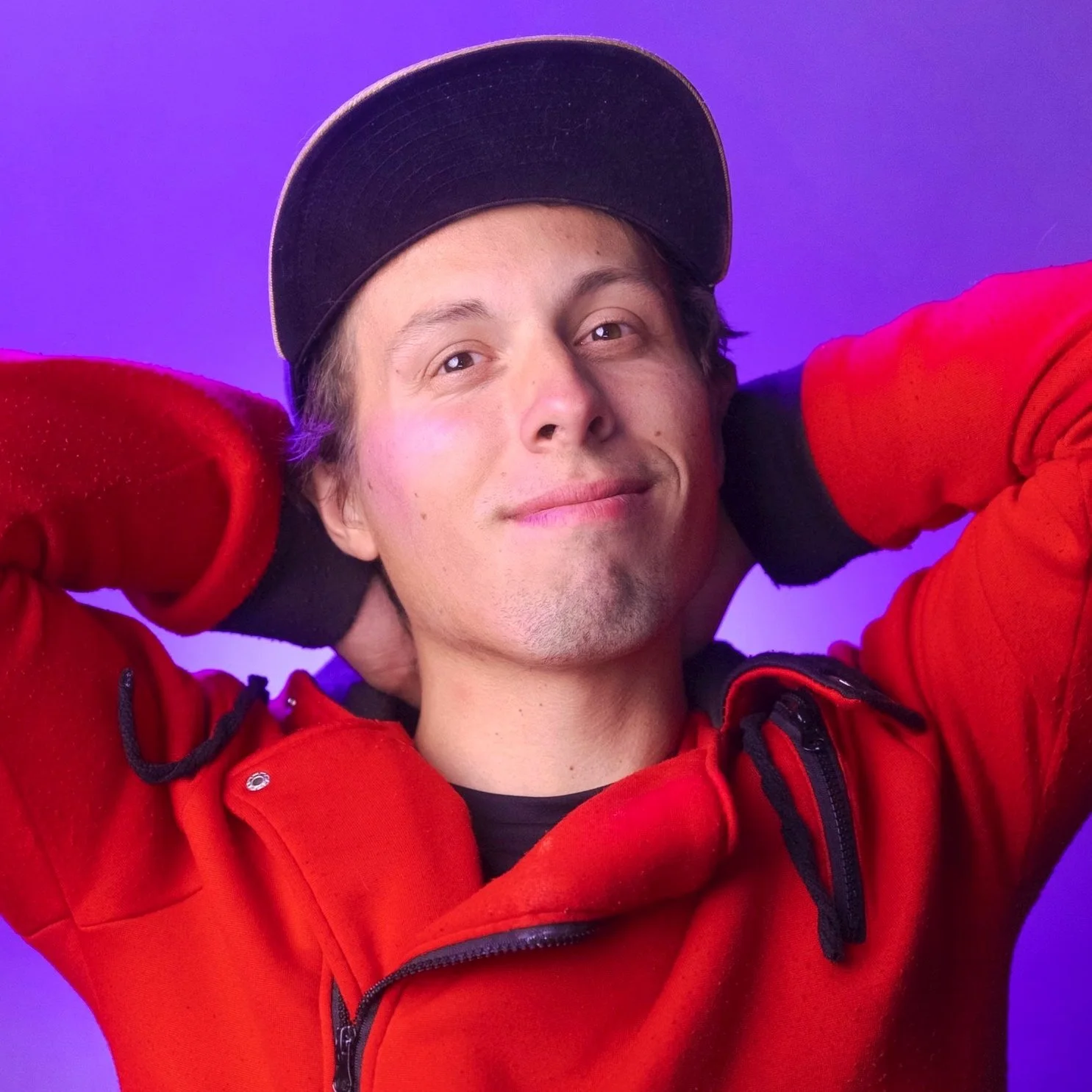 A young man with glasses smiling, wearing a black cap with a 'Gyro Studios' logo, a black T-shirt with a red and yellow graphic, and a backpack against a purple background.