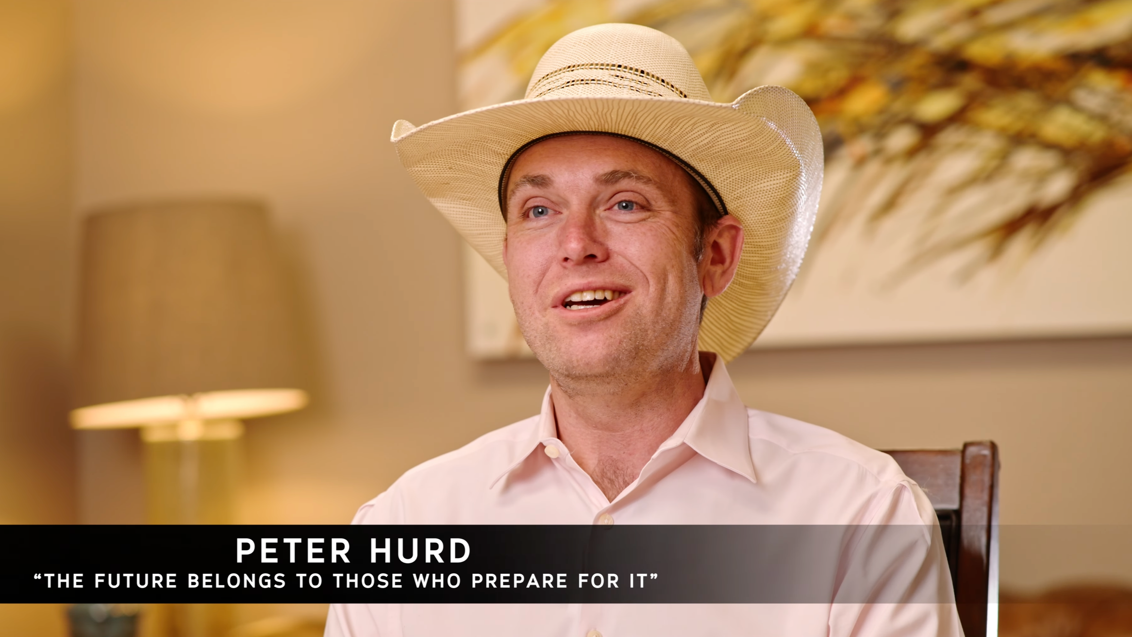 A man wearing a beige cowboy hat and a light pink dress shirt is sitting indoors, smiling. There is a lampshade and a floral painting in the background. The caption reads: Peter Hurd, 'The future belongs to those who prepare for it.'