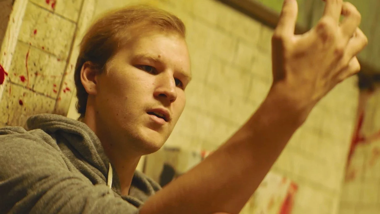 A young man with short red hair, wearing a grey hoodie, sits against a wall with bloody markings. His face appears distressed and he is reaching out with his hand.