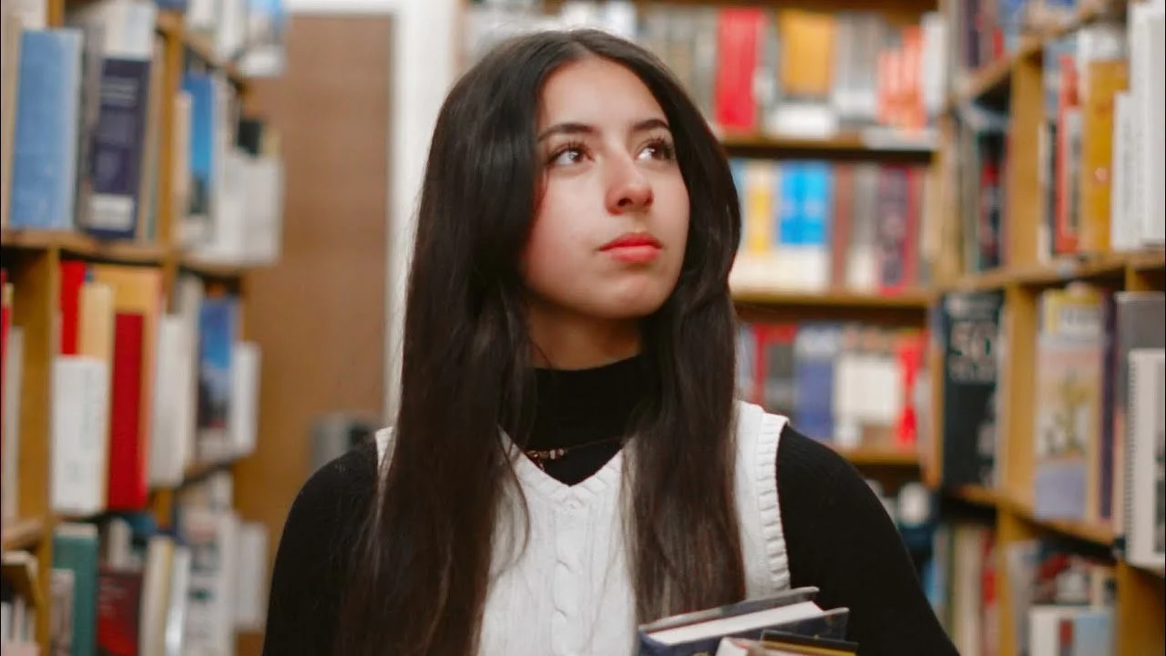 A young woman in a library, holding books, looking up thoughtfully.