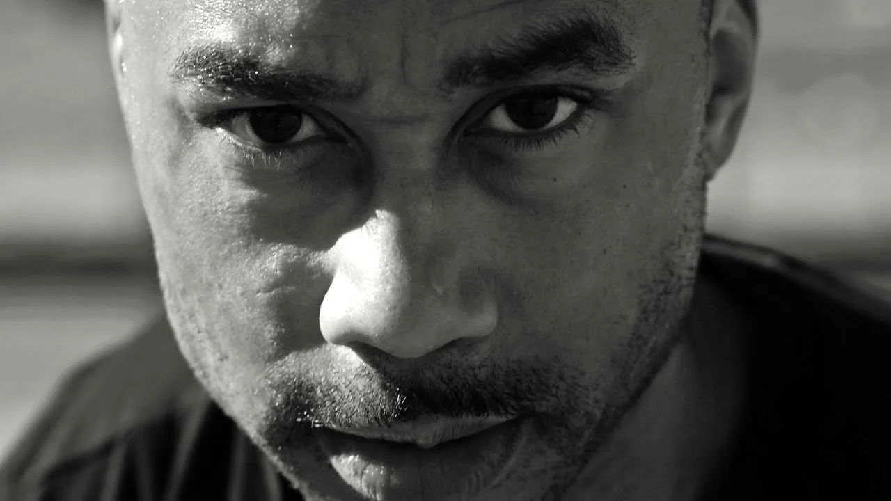 Close-up black-and-white photo of a man with intense eyes, slightly sweaty face, and facial hair, looking directly at the camera.