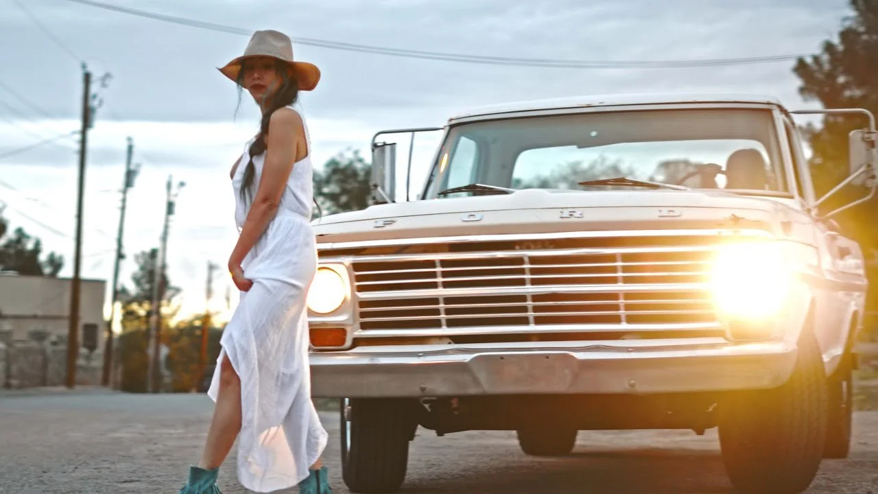 A woman in a white dress and a wide-brimmed hat stands next to a vintage Ford truck with its headlights on during sunset.