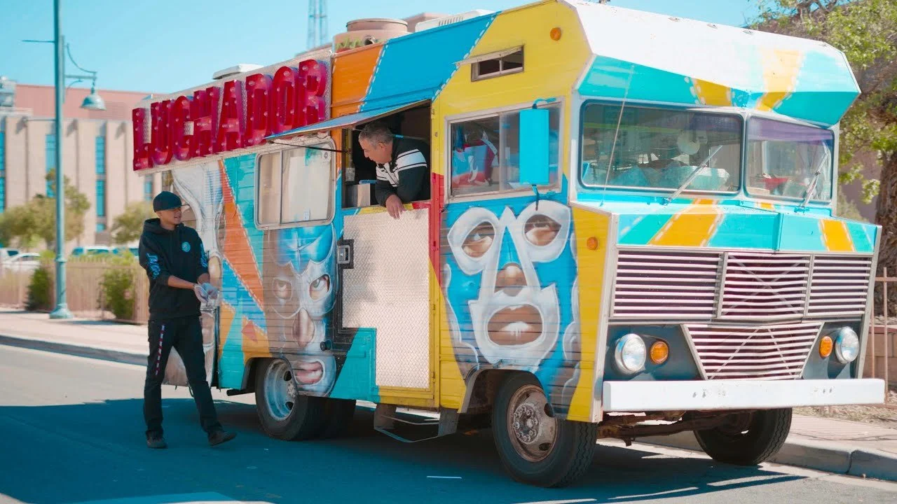 A colorful food truck with fitness-themed artwork parked on the street. Two people are interacting with the truck; one is inside the truck leaning out of the window, and the other, standing outside, is wearing a black hoodie and a beanie.