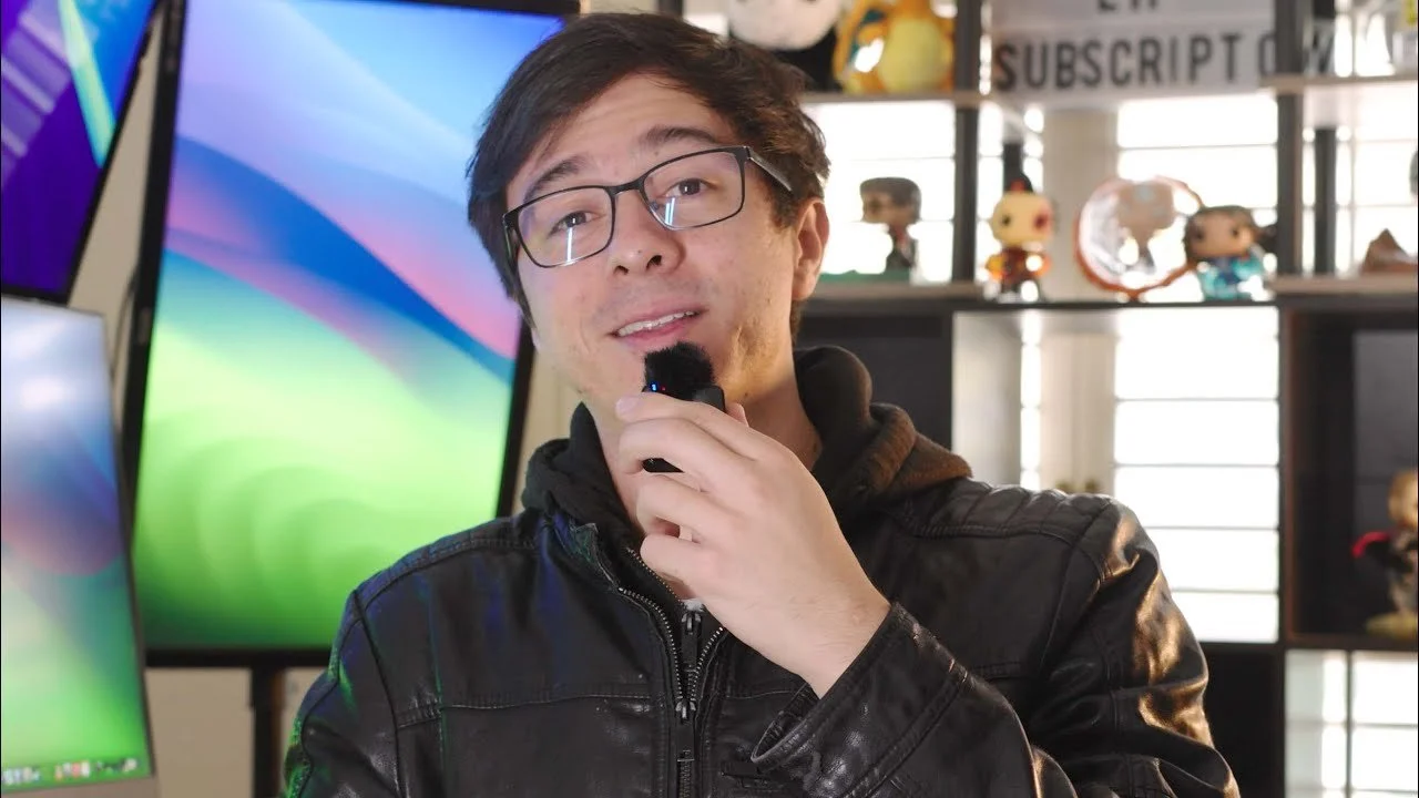 A young man with glasses and dark hair, wearing a black leather jacket, holding a microphone in his hand, in a room with multiple monitors and figurines in the background.