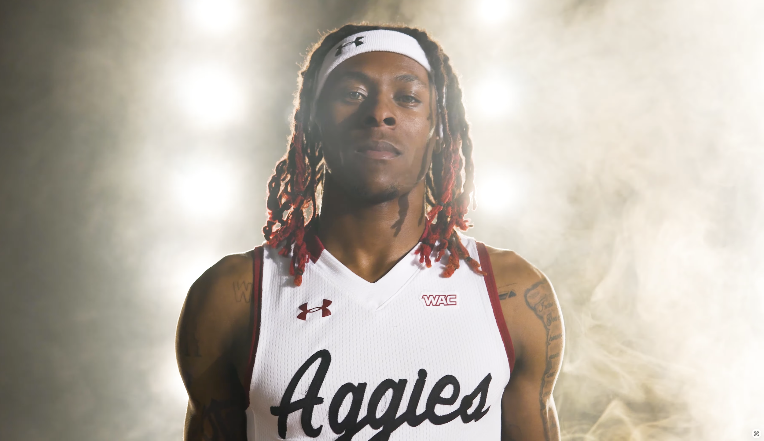 A female athlete with red and black dreadlocks wearing a white basketball jersey with red accents, standing in front of bright lights and smoke, looking into the camera.