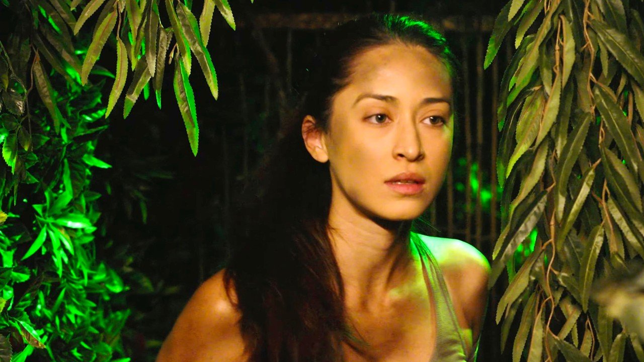 A woman with long dark hair stands among green tropical plants at night, lit by green lights.