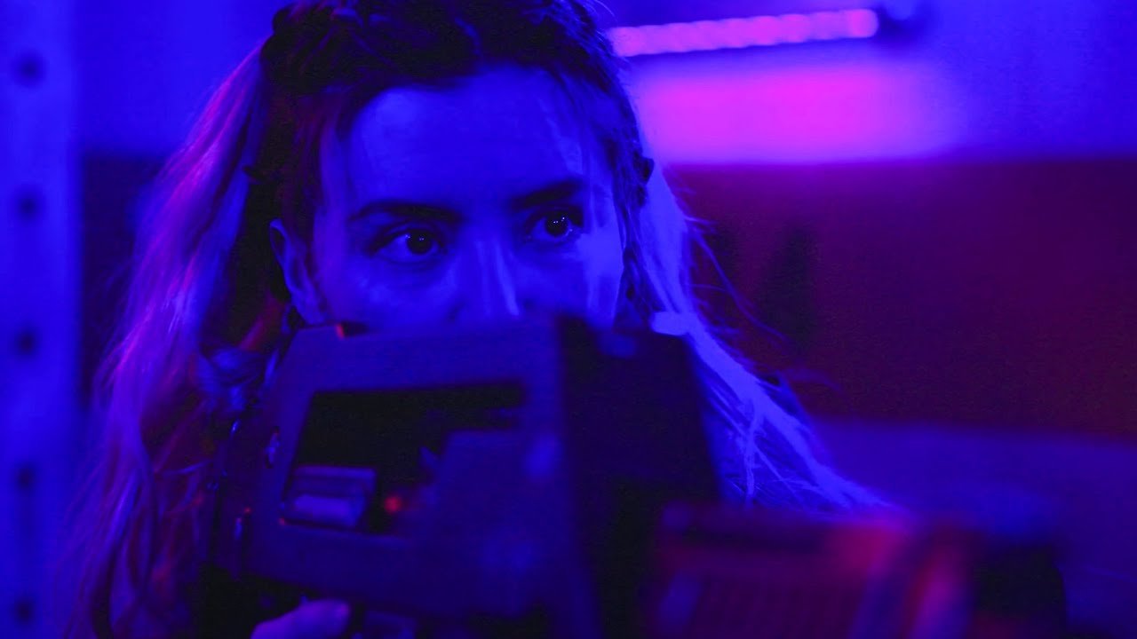 A woman with long hair holding a gun in a dimly lit room with purple and blue neon lighting.