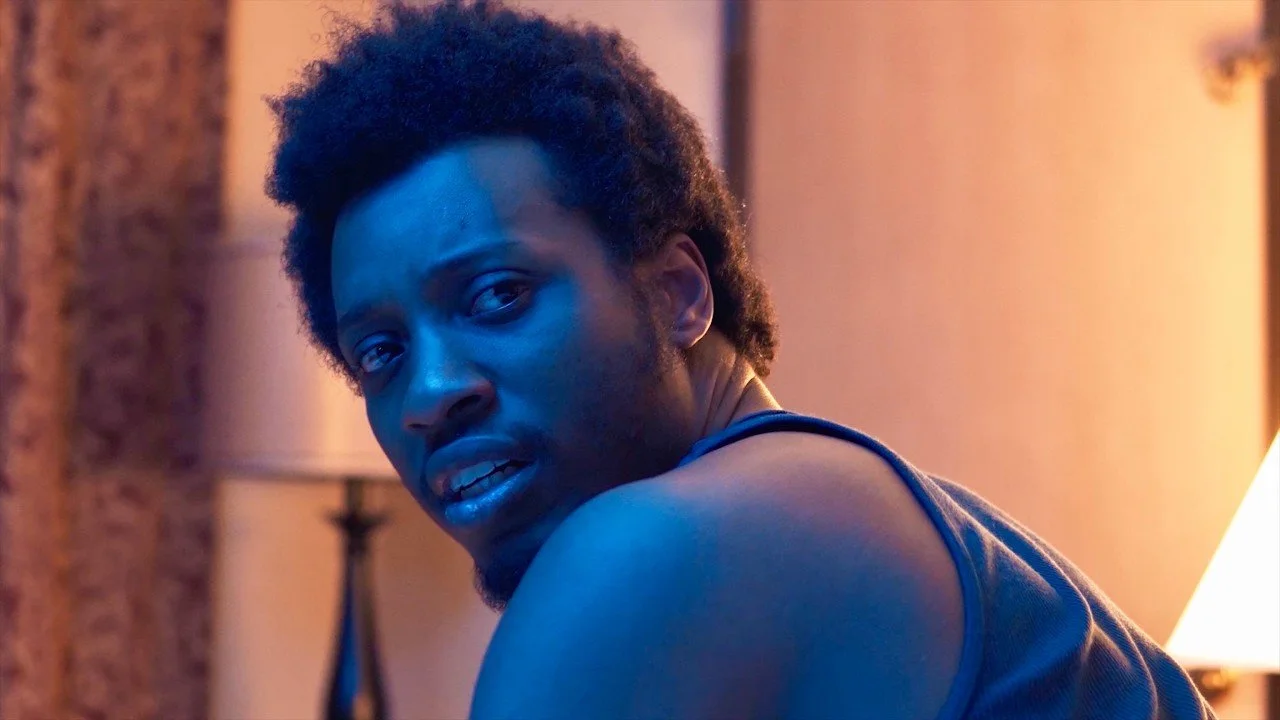 A young man with curly hair and a beard looking over his shoulder at the camera, illuminated by blue and warm lighting in a cozy indoor setting.