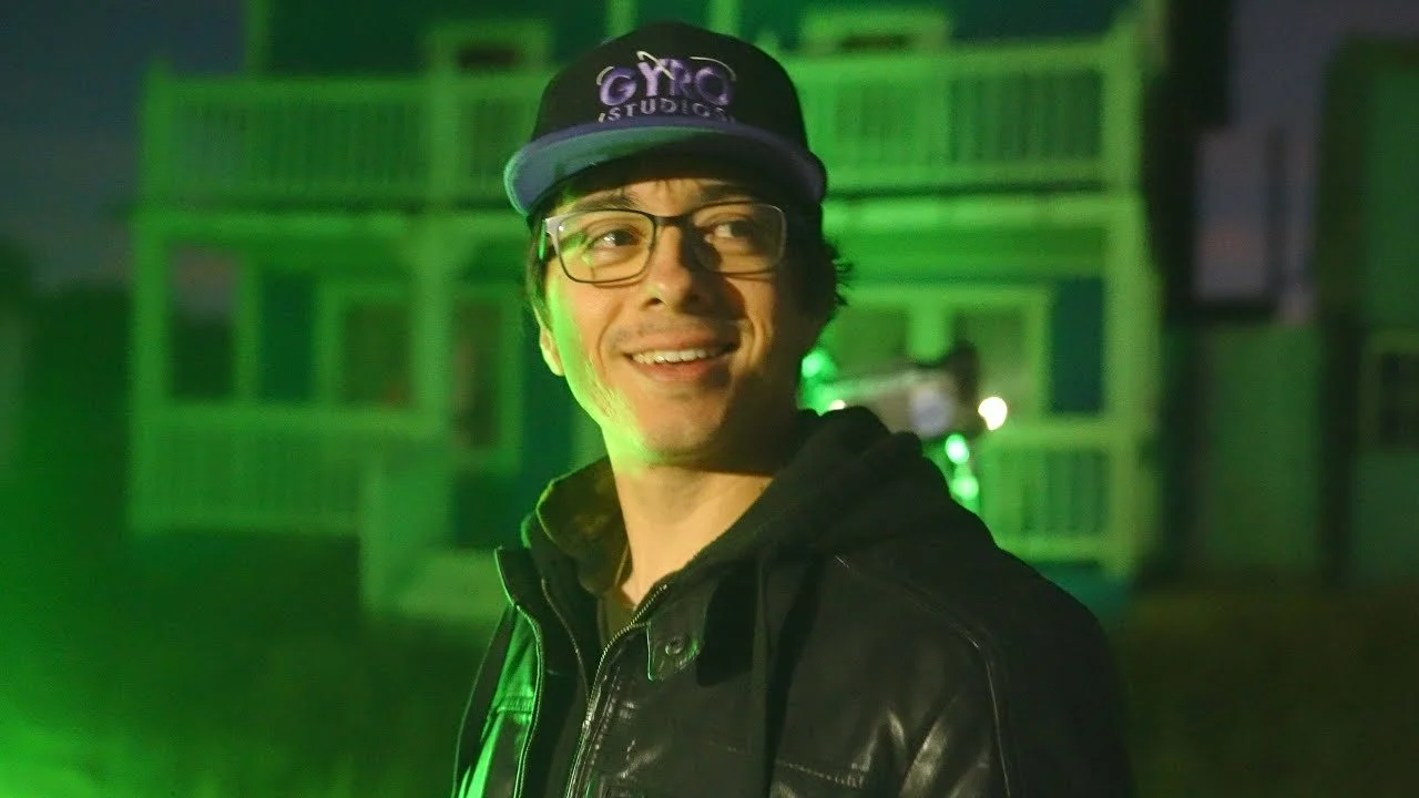 A smiling young man wearing glasses, a black baseball cap with purple text, and a black leather jacket, illuminated by green stage lighting in front of a wooden building.