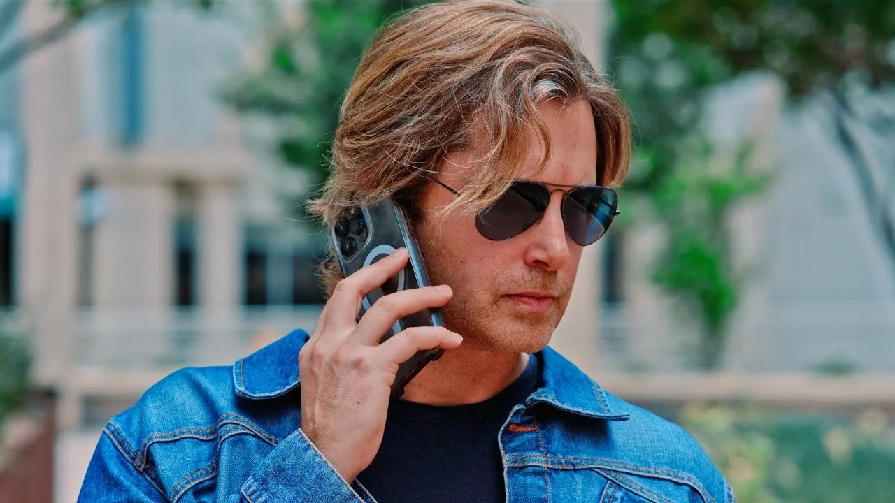 A man with shoulder-length wavy brown hair wearing sunglasses and a denim jacket is talking on a cell phone outside.