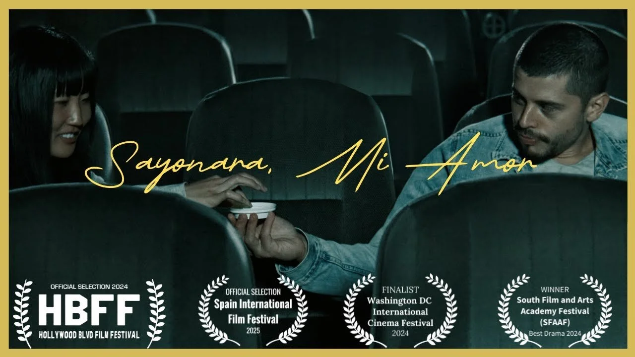 A woman and a man sitting in a theater, with the woman handing the man a small item, likely a plate, over the back of a seat. The scene appears intimate, with the text "Sayonda, Mi Amor" overlayed. The image also displays several film festival awards and nominations at the bottom.