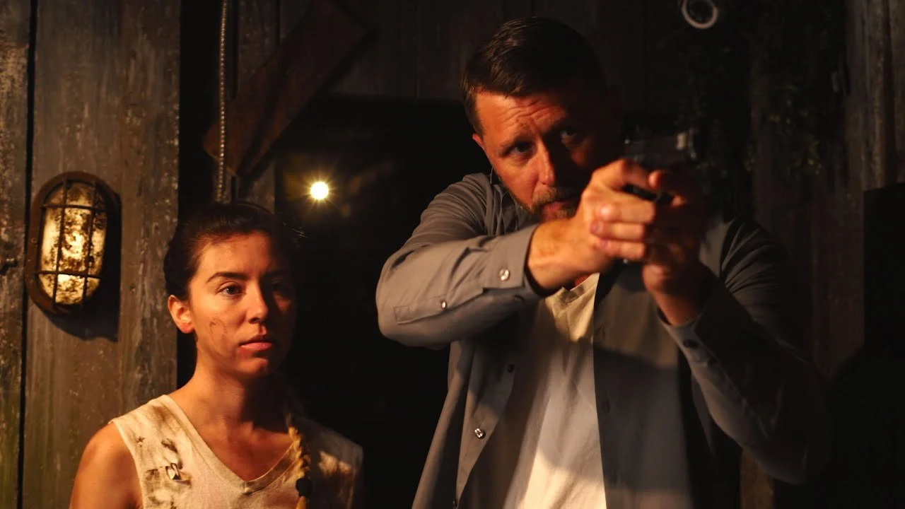A man aiming a handgun in a dark room with a woman standing nearby, looking tense.