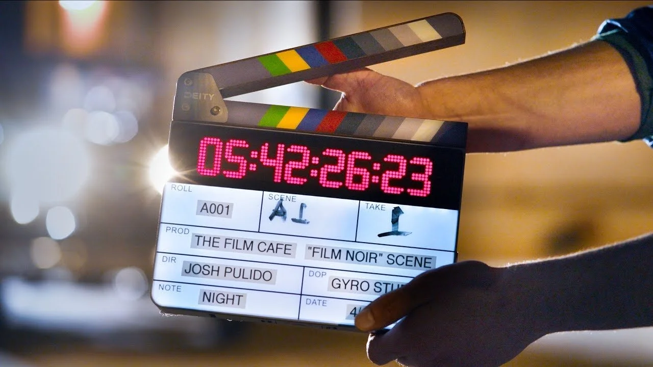 A film production slate displaying details of a scene, including roll number, scene, take, production title, director, note, and date, with a countdown timer and a person's arm holding it.