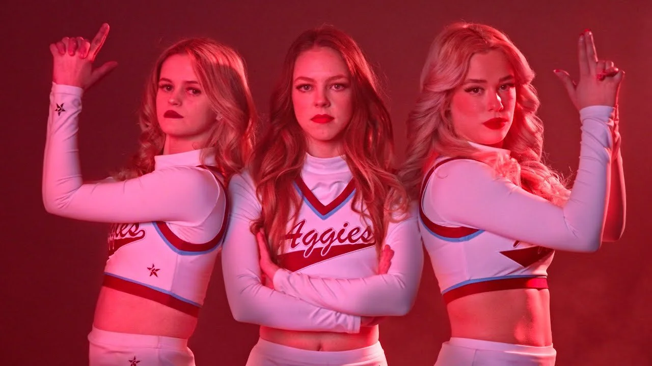 Three women in cheerleading uniforms with the word 'Aggies' on the front, posing confidently with arms raised in a formation against a red background.