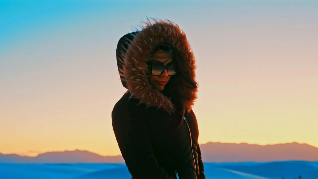 A woman wearing a black winter coat with a fur-lined hood, sunglasses, and a beanie hat, standing outdoors during sunset with mountains and a colorful sky in the background.