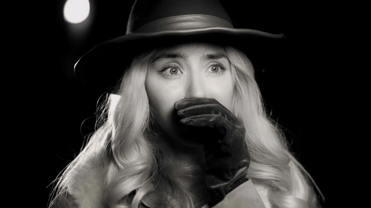 A woman with long wavy hair wearing a hat and glove, covering her mouth with her hand, in black and white.