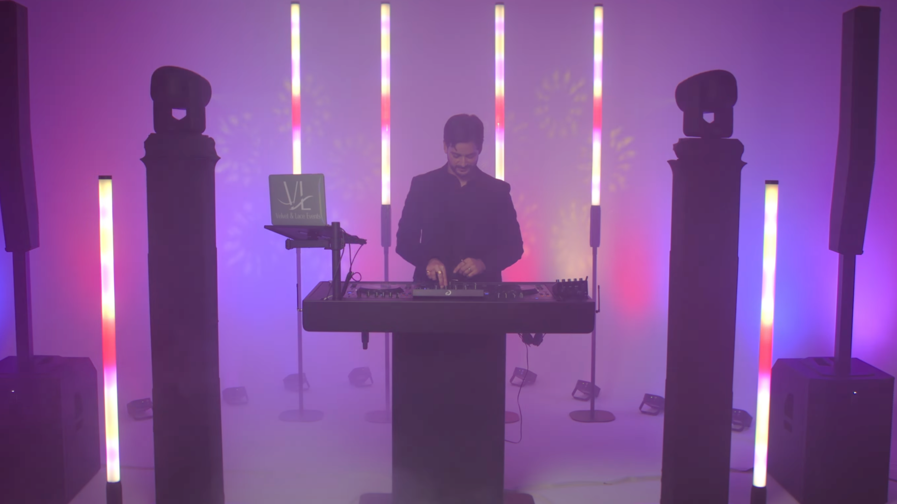A DJ performing in a club with colorful vertical LED lights, standing behind a DJ booth, with speakers and a laptop displaying 'Sly' and 'Mikael & Lace Events' on the screen.