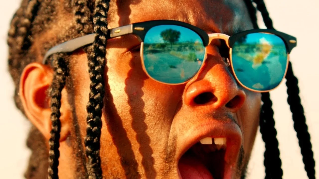 Close-up of a man with braided hair wearing sunglasses, with reflections of a landscape in the lenses.