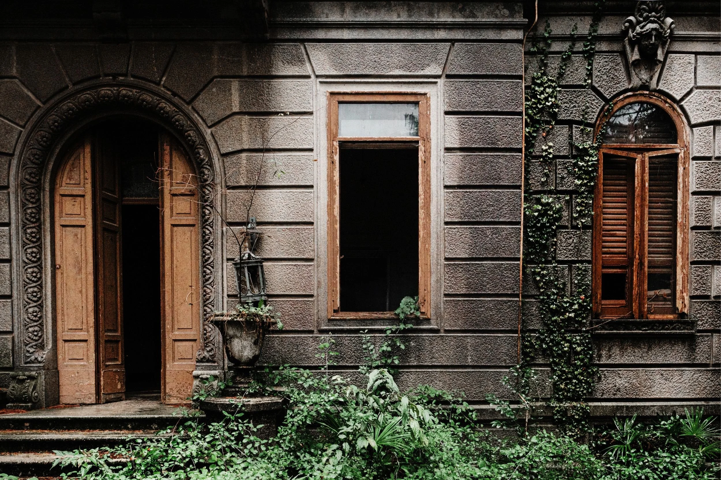 The façade of an abandoned villa, ivy over the stone and the door ajar