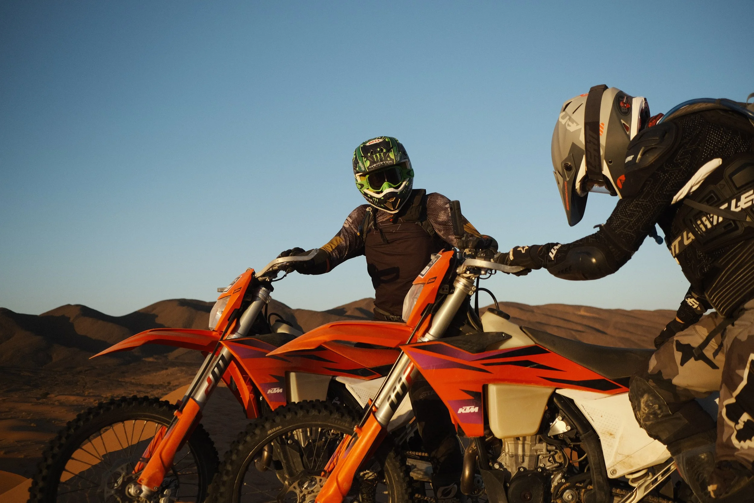 Two riders on dirt bikes in the desert, one reaching out to the other
