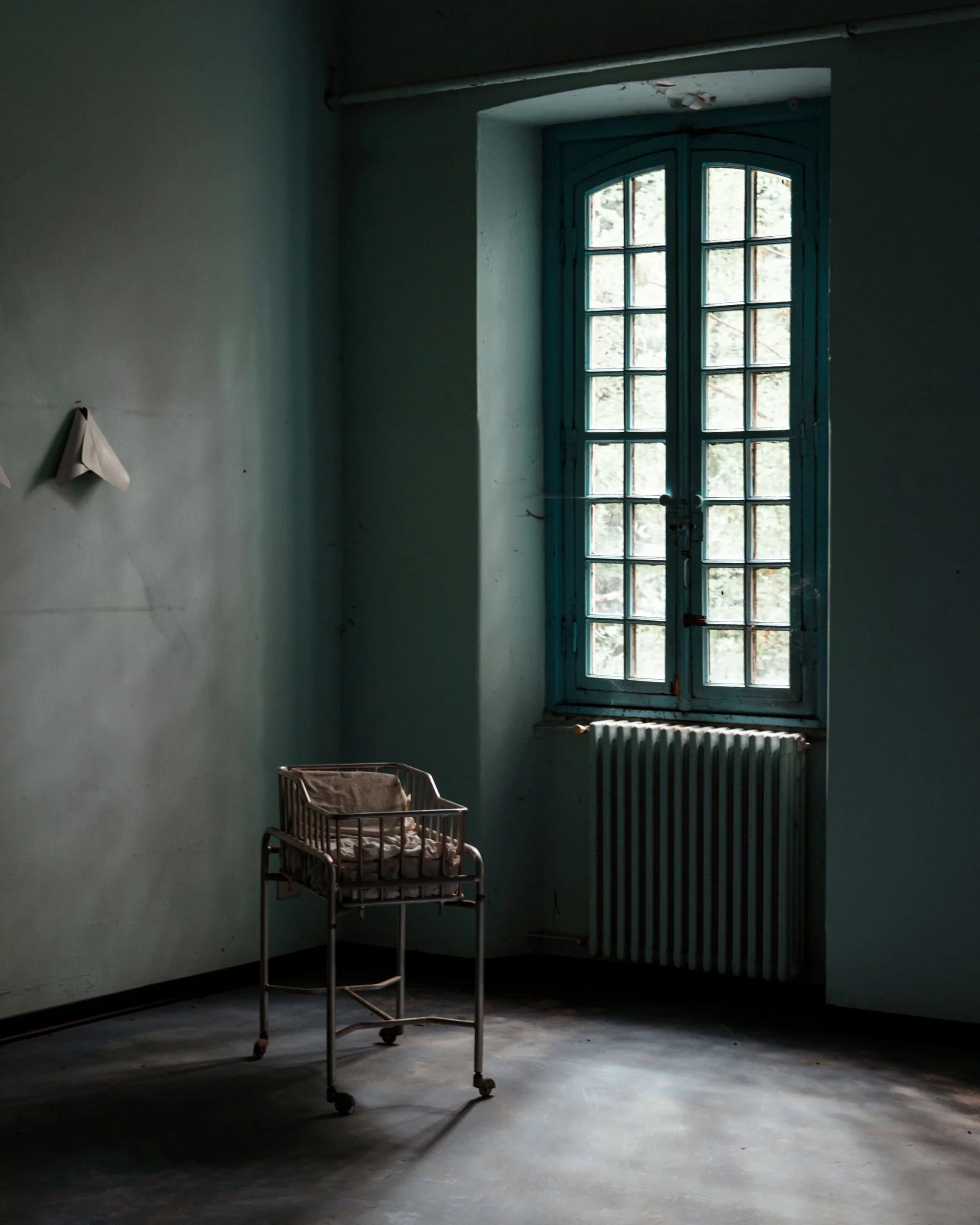 A single crib by a tall window in a green-walled room of an abandoned asylum