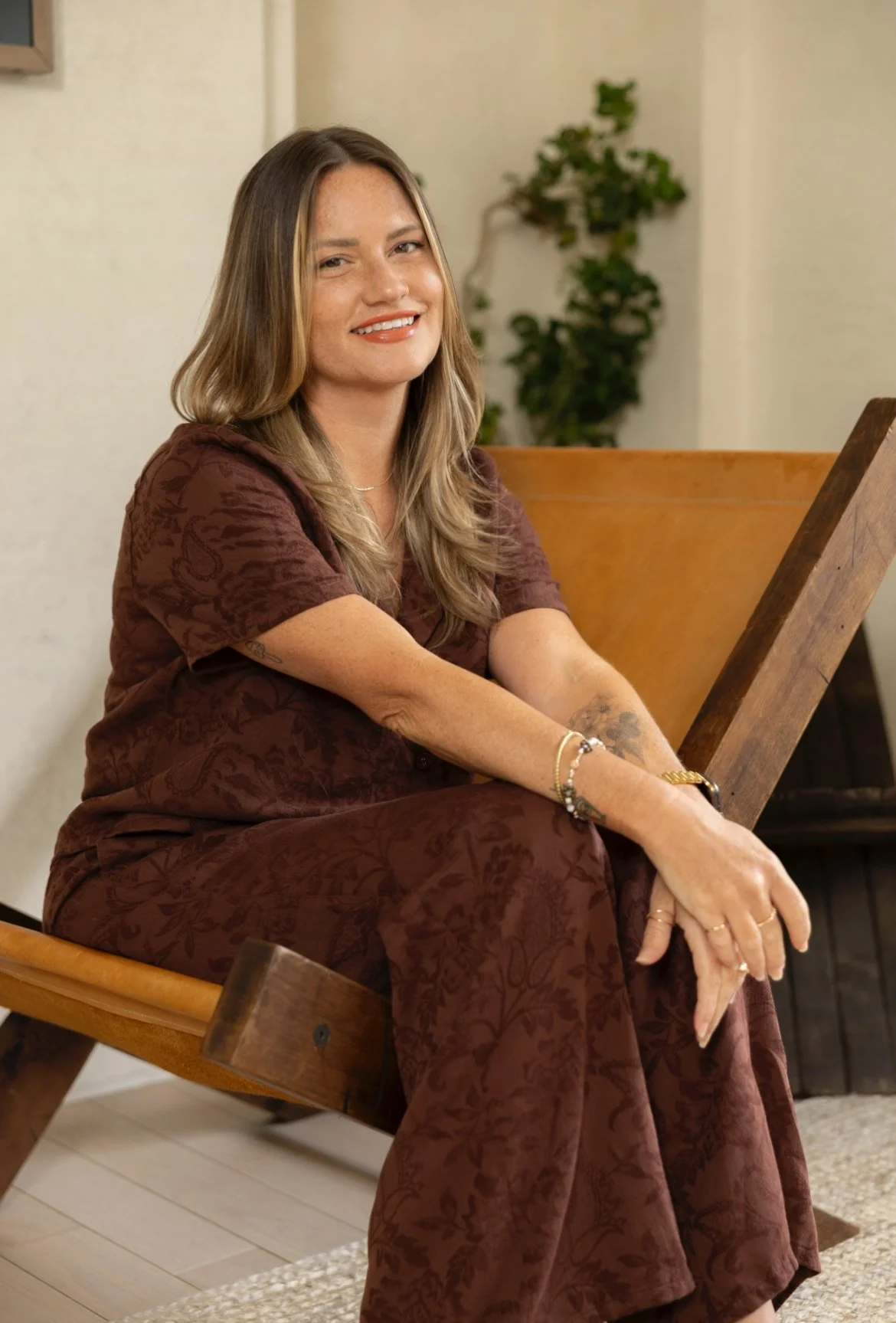 A woman with long, wavy, brown hair and fair skin sitting on a wooden bench indoors. She is wearing a matching brown patterned outfit and several bracelets.