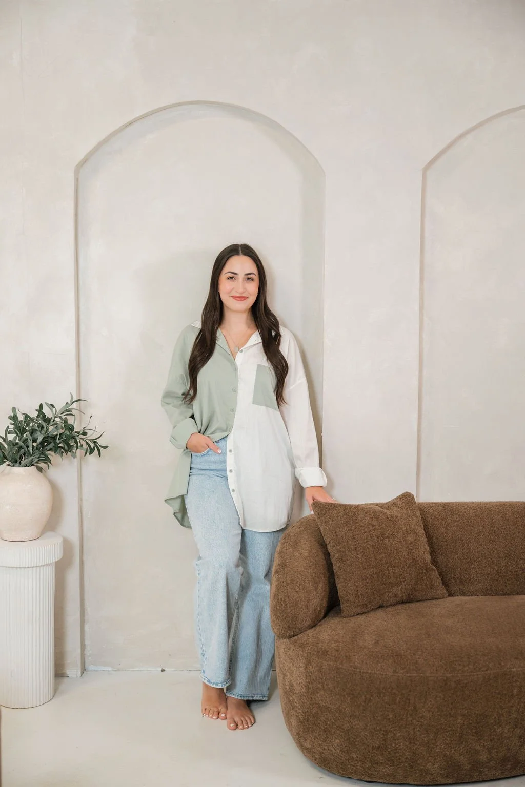 A woman with long dark hair, wearing a color-blocked shirt, and light-colored jeans, standing barefoot indoors, with a brown couch and a potted plant nearby.