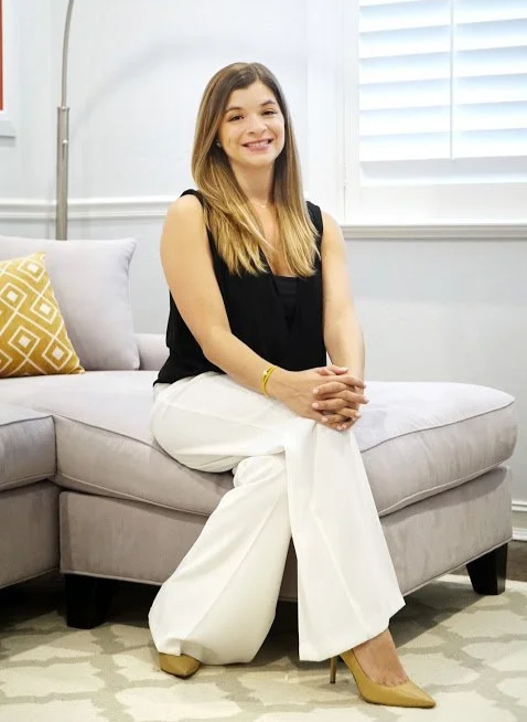A woman with long, light brown hair sitting on a light gray sofa in a bright room with white blinds. She is wearing a black sleeveless top, white wide-leg pants, and beige high heels, smiling at the camera.