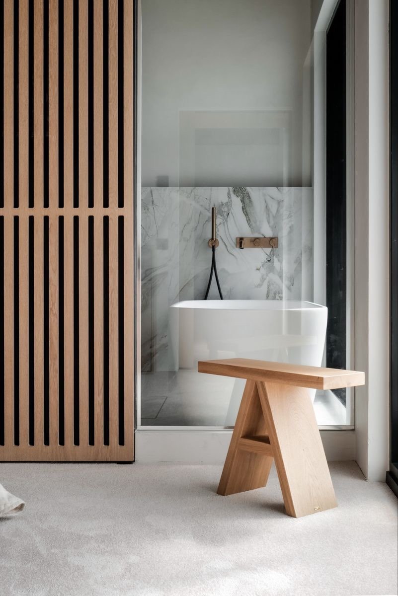 Modern bathroom with a freestanding bathtub, marble wall, black showerhead, and a wooden stool near a glass wall.