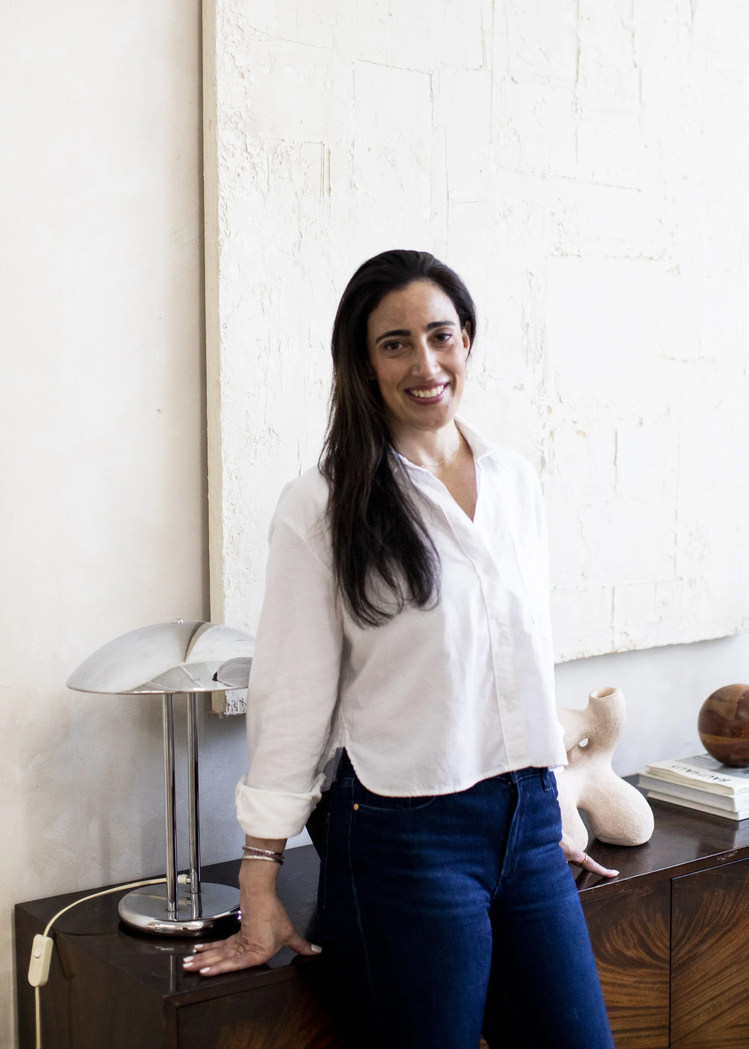 A woman with long dark hair, wearing a white blouse and dark jeans, standing next to a sideboard with decorative objects and artwork in a bright room.