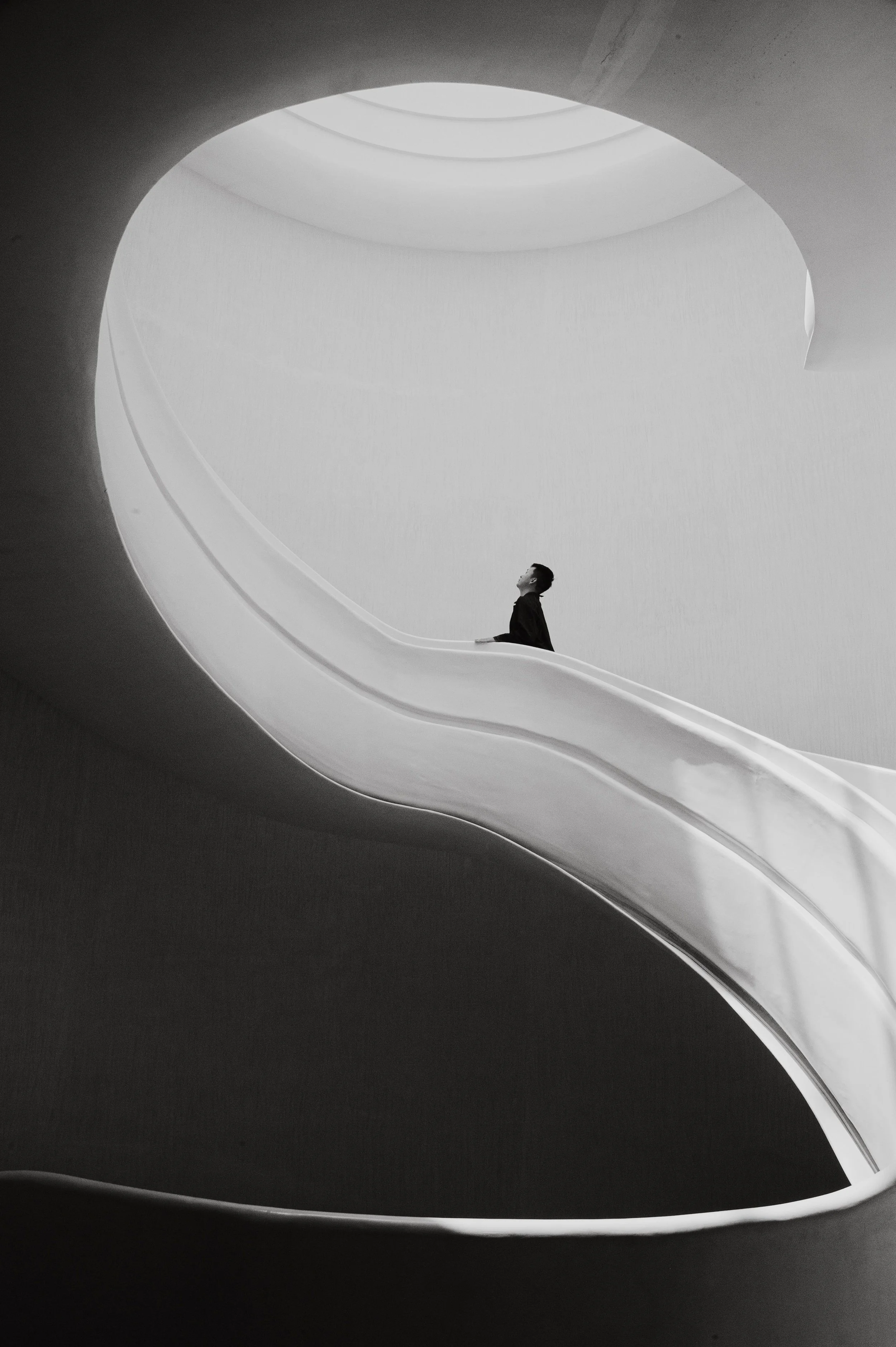 A person in dark clothing standing on curved staircase within modern, circular architectural space, looking upward.