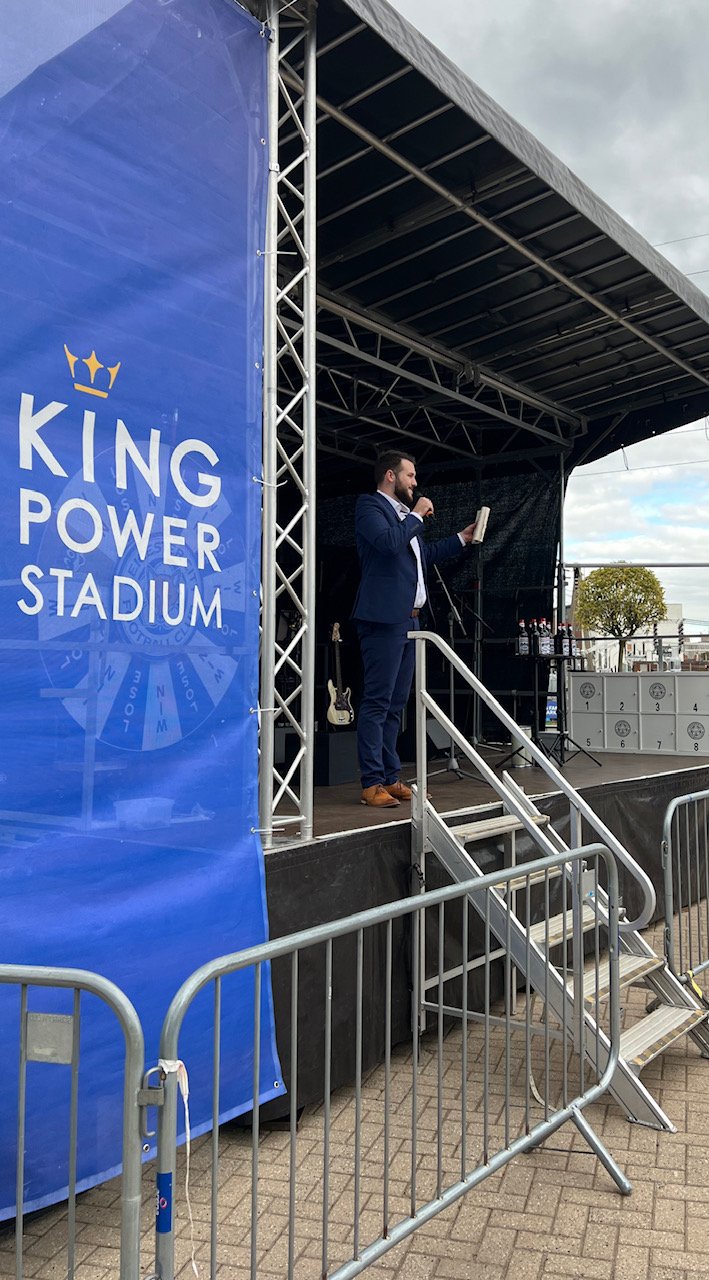 Man in a suit speaking on an outdoor stage near King Power Stadium sign, with microphones and equipment in the background.