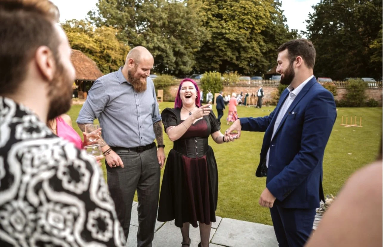 Group of people laughing and talking at an outdoor event; one person with pink hair, another with a beard, one in a suit; trees and lawn in the background.