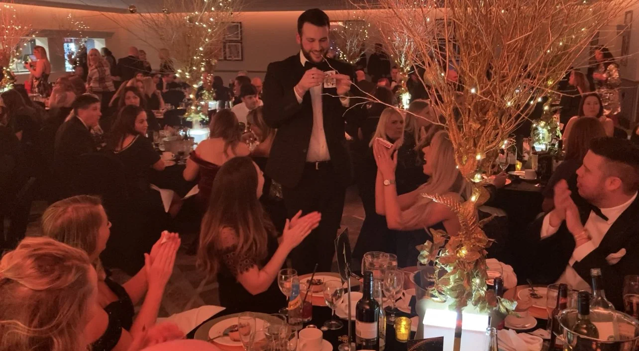 A formal event with a man standing and smiling, performing a magic trick for a group of seated guests. The table is elegantly decorated with glasses, bottles, and a golden plant centerpiece. The guests are dressed in formal attire, clapping and watching attentively.