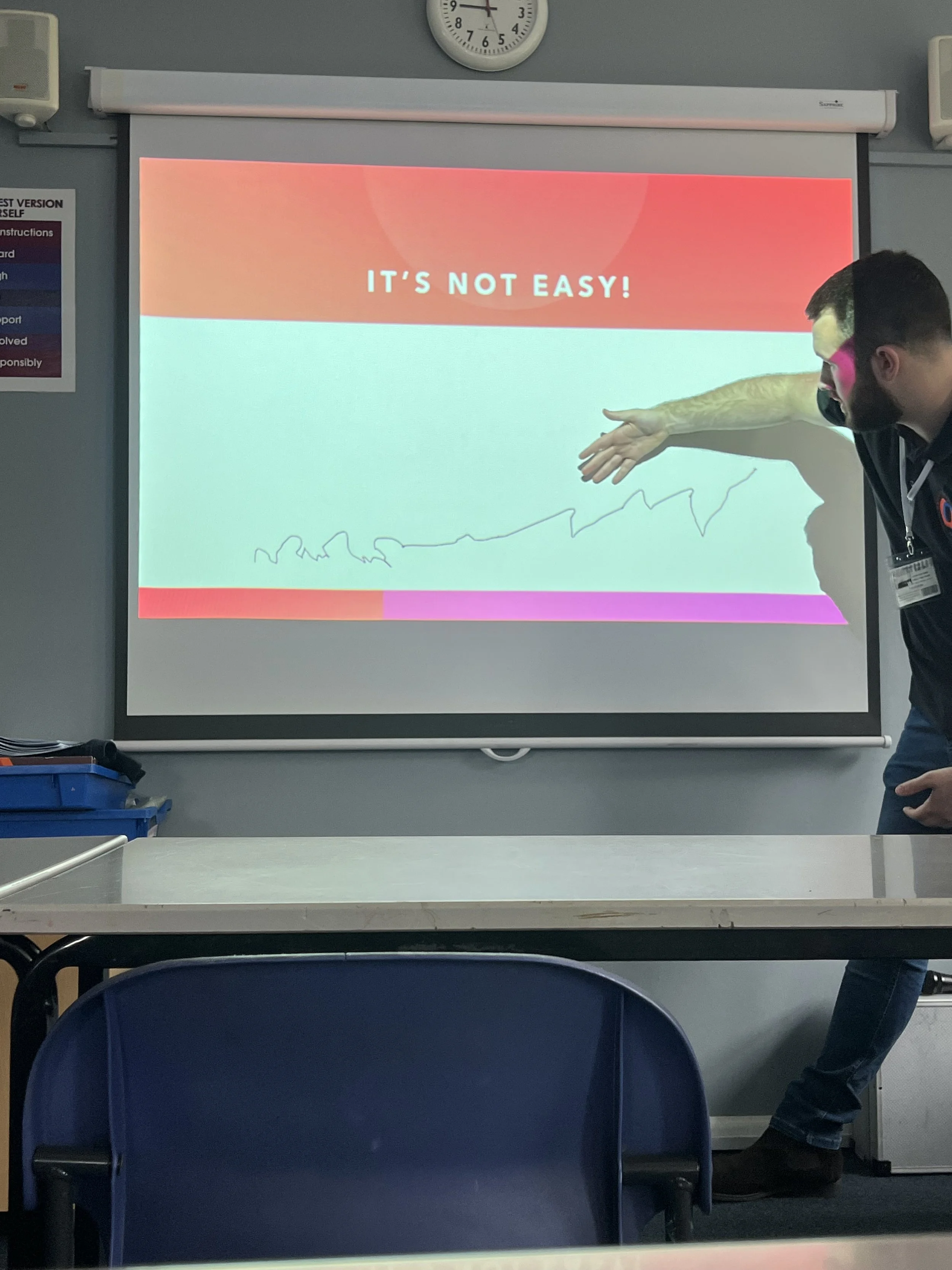 Person presenting a slide that says 'It's Not Easy!' with a graph-like scribble on a classroom projection screen.