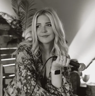 A woman with long blonde hair holding a microphone, smiling softly, sitting indoors near a plant.