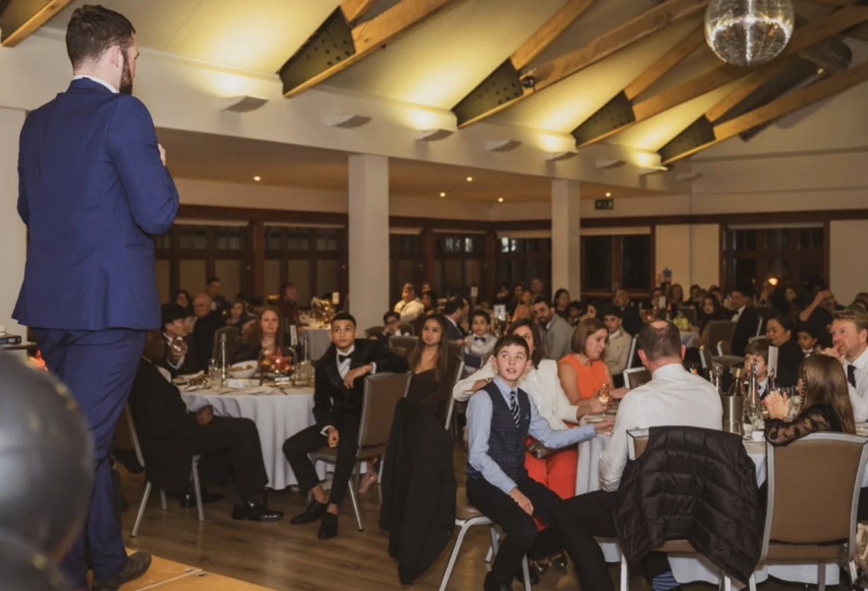 A man in a blue suit speaks to an audience seated at round tables in a banquet hall. The room is well-lit with a modern wood beam ceiling, and attendees are dressed in formal attire, attentively listening to the speaker. A disco ball hangs from the ceiling.