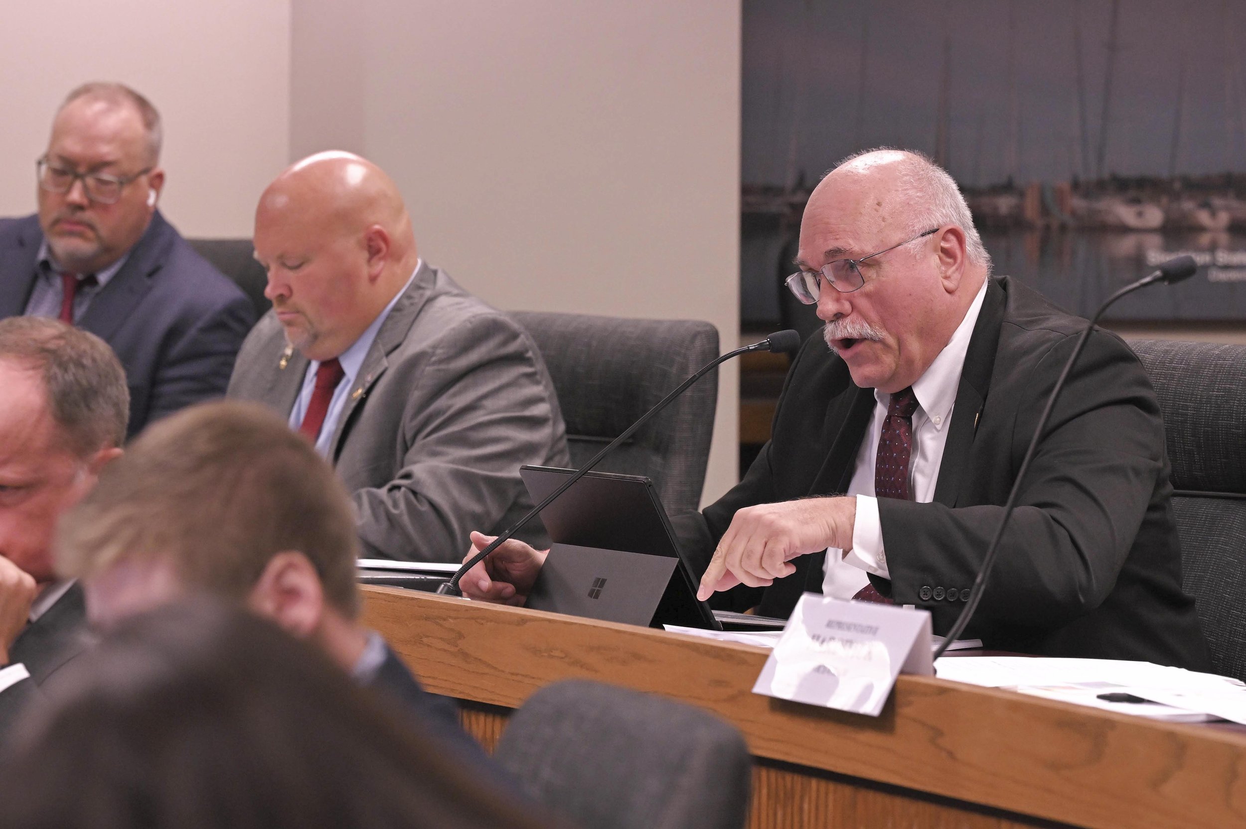 A man with glasses, a white mustache, and a bald head, wearing a black suit and a white shirt, is speaking into a microphone while seated at a conference table with a tablet in front of him. Other men in suits are also seated at the table, some with 