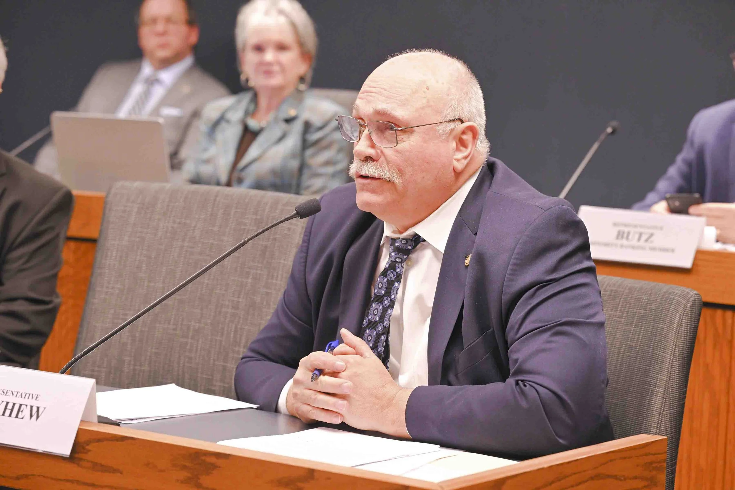 A man with white hair, glasses, and a mustache, dressed in a navy suit and patterned tie, is speaking into a microphone at a formal meeting or hearing. He is sitting at a desk with papers in front of him, and there are others seated behind him in the