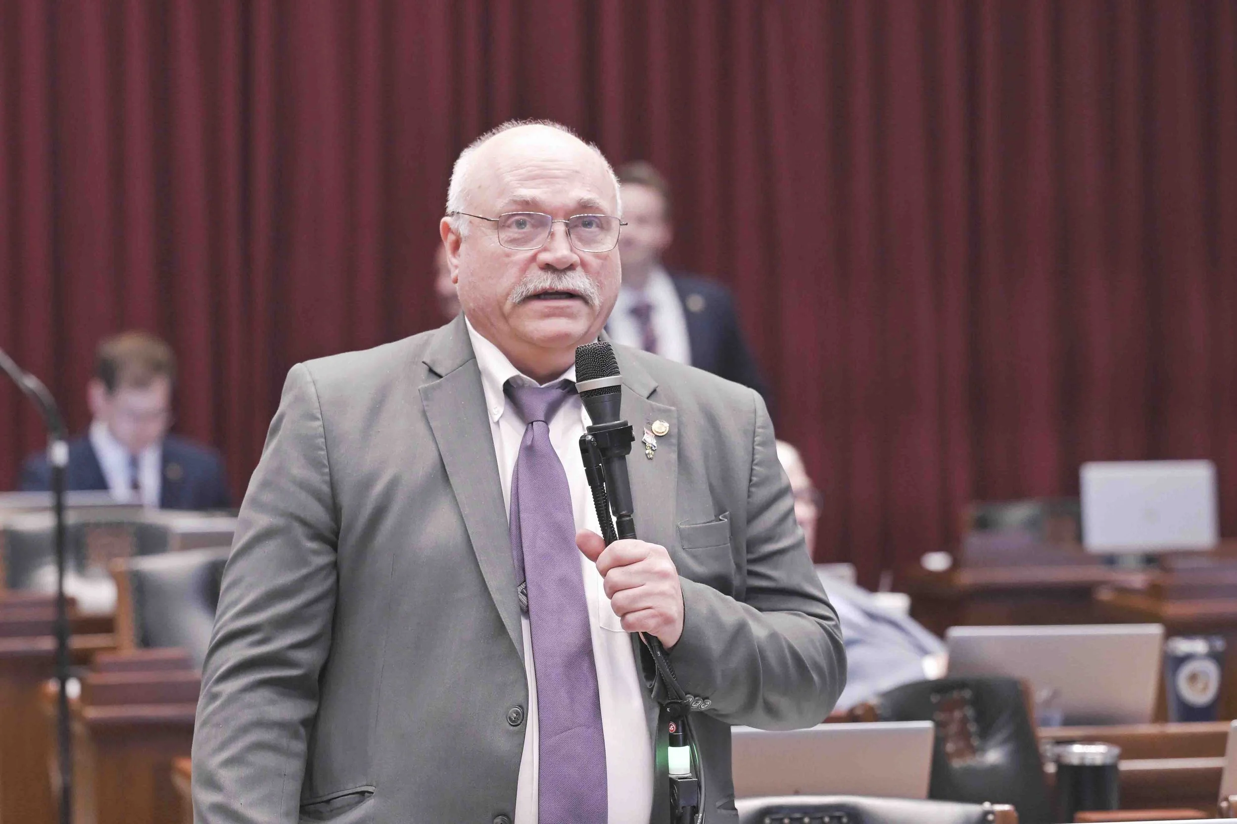A man with glasses, a gray suit, and a purple tie speaking into a microphone in a formal setting with red curtains in the background.